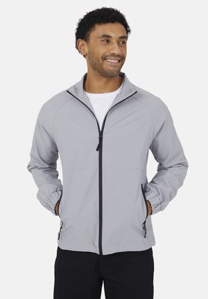 Uśmiechnięty mężczyzna w jasnoszarej kurtce na zamek z elastycznymi mankietami i czarnymi detalami zamka, stojący z rękami w kieszeniach.