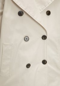 Manteau croisé beige clair avec des boutons ronds foncés et une poche verticale fendue sur le côté gauche.