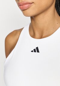 Camiseta deportiva blanca sin mangas con escote redondo. Presenta un logo negro de Adidas en el lado superior izquierdo.