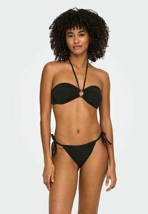 Woman with curly hair wearing a black strapless bikini top with ring detail and matching tie-side bikini bottoms, standing against a plain background.