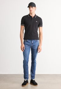 Fekete póló fehér gallér díszítéssel, kék farmer, fekete sportcipő és fekete sapka. Slim fit fazon CK logóval a mellkason.