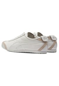 Paire de baskets basses blanches et beiges avec lacets sur le devant et marque "Tiger" sur les languettes de talon, visibles de dos et de côté.