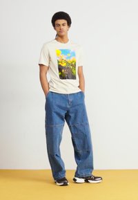 Tricou bej cu mânecă scurtă, având un grafic floral colorat pe față, asortat cu blugi evazați de un albastru deschis și adidași negri.