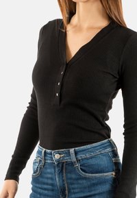 Haut noir à manches longues côtelé avec un décolleté en V et trois boutons dorés. Associé à un jean en denim bleu légèrement décoloré avec des poches avant.