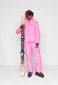 Chaqueta y pantalones de esquí rosas, material impermeable, diseño ajustado. Esquiador sosteniendo esquís con fijaciones, llevando una máscara de esquí negra y gafas.