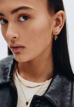 Goldene, herzförmige Ohrringe hängen an den Ohren des Models. Sie trägt eine schwarze Lederjacke über einem weißen Pullover und hat mehrere goldene Halsketten übereinander.