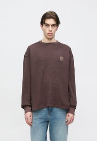 Rudas tekstūruotas ilgomis rankovėmis džemperis su apvalia iškirpte ir ribotais rankogaliais, su mažu oranžiniu logotipo pleistru ant krūtinės.