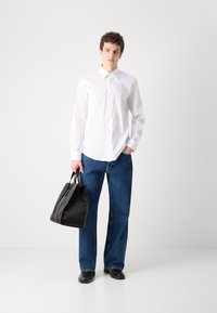 Bela srajca z gumbi, modri jeans in črna usnjena torba z teksturiranimi akcenti. Moški stoji samozavestno, roke v žepih.