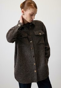 Brązowa kurtka koszulowa w kratę houndstooth wykonana z grubego materiału, z ozdobnymi złotymi guzikami i dwoma kieszeniami na piersi. Luźny, swobodny fason.