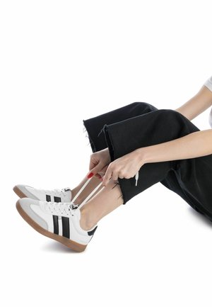 Sneakers bianchi con strisce nere, dettagli in suede e suole in gomma, abbinati a pantaloni neri a vita alta, persona che allaccia le scarpe.