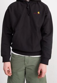 Schwarzer Kapuzenpullover mit einer vorderen Tasche, der ein kleines rotes und gelbes Logo trägt. Weicher Stoff, gerippte Bündchen und Saum. Kombiniert mit grünen Hosen.