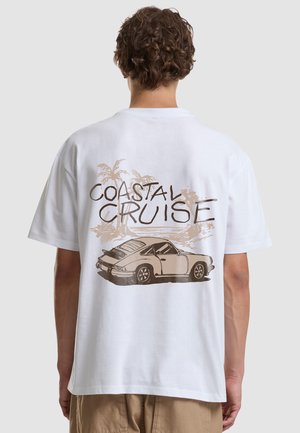 Inimene, kellel on lokkis juuksed ja valge t-särk, näitab vintage autot ja palme, millel on seljal tekst "Coastal Cruise".
