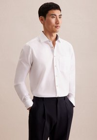 Camicia bianca con maniche lunghe, dotata di taschino sul petto e dettagli decorativi sul colletto, abbinata a pantaloni scuri. Tessuto dalla trama liscia.