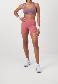 Sujetador deportivo morado claro y pantalones cortos altos rosas de tela elástica, combinados con zapatillas deportivas blancas y muñequeras.