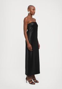 Vestido negro de satén sin tirantes con diseño hasta el suelo. Textura suave, silueta ajustada, combinado con sandalias de tacón alto negras con tiras.