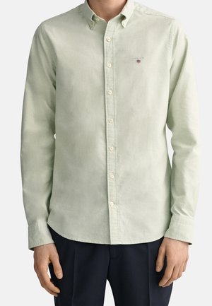 Uomo che indossa una camicia a maniche lunghe verde chiaro con bottoni e un piccolo logo sul petto, abbinata a pantaloni scuri, in piedi con le braccia rilassate.