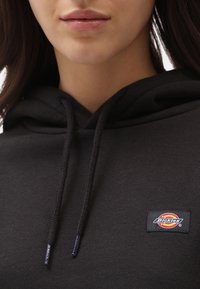 Sweat à capuche noir avec cordons, arborant un petit écusson de logo coloré sur la poitrine. Le tissu est lisse, avec une texture douce.