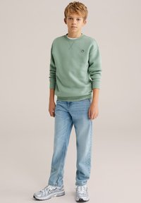 Blond chlapec oblečený v šalviovo zelenej mikine, svetlomodrých džínsoch a strieborných teniskách Nike, stojaci pred jednoduchým béžovým pozadím.