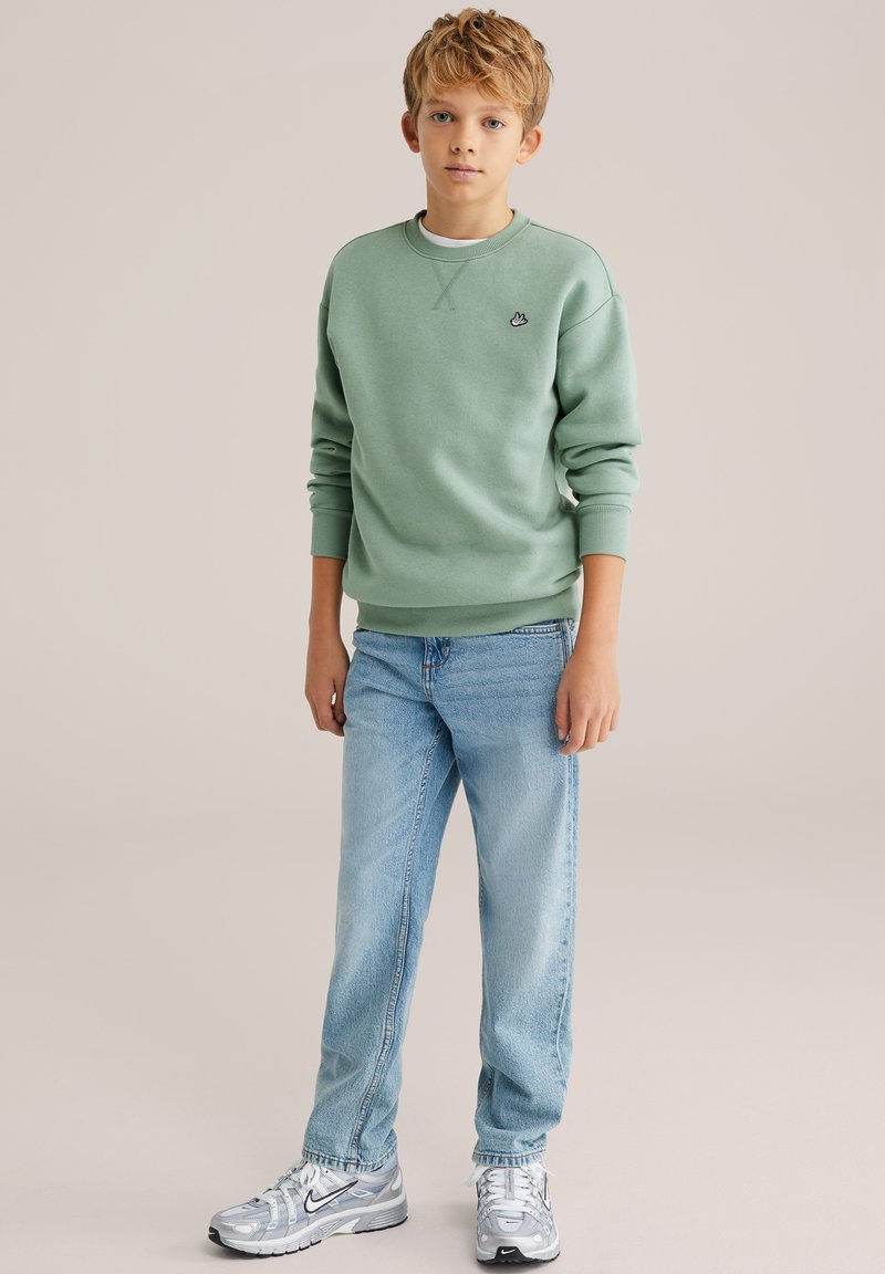 Blond chlapec oblečený v šalviovo zelenej mikine, svetlomodrých džínsoch a strieborných teniskách Nike, stojaci pred jednoduchým béžovým pozadím.