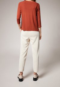 Top arancione a maniche lunghe abbinato a pantaloni bianchi testurizzati e scarpe beige con suole nere spesse. Design semplice e minimalista.
