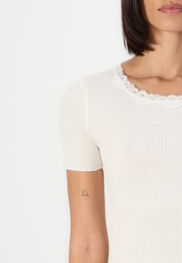Ribbed valkoinen lyhyet hihat -paita, jossa on aallotettu pitsikaulus, sileä pinta ja vartalonmyötäinen malli. Huomaamaton tatuointi näkyy kädessä.
