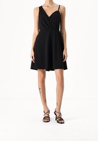 Musta hihaton mekko, jossa on kietaisumuotoilu ja levenevä hame, valmistettu sileästä kankaasta. Yhdistetty mustiin remmikenkiin.