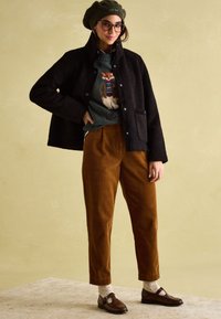 Young woman wearing green beret, black fleece jacket, green sweater with fox design, brown corduroy pants, white socks, and brown loafers, standing.