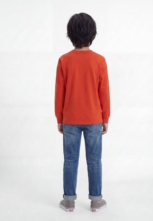 Enfant aux cheveux foncés, debout de dos, portant un t-shirt à manches longues orange, un jean bleu roulé et des baskets grises sur fond blanc.