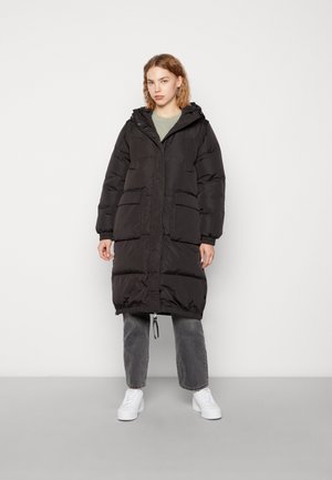 Svart puffjacka med huva, med en framsida som har dragkedja, två stora fickor, elastiska ärmar och dragsko i nederkant, bärs över grå jeans och vita sneakers.