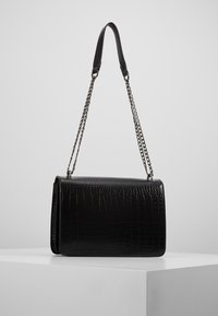 Schwarze Handtasche aus Kunstleder mit Krokodil-Textur, strukturierter Form und abnehmbarem silbernen Kettenriemen. Glatte Akzente vervollständigen das Design.