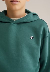 Vihreä huppari pehmeästä kankaasta, jossa on kengurupussi ja pieni brodeerattu lintulogot vasemmassa rinnassa.