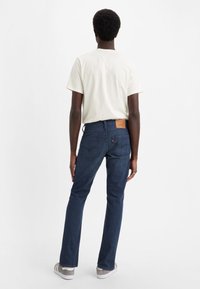 Jeans in denim blu scuro con un taglio dritto classico, dotati di una patta in pelle marrone e tasche posteriori. Abbinati a una maglietta di un leggero color crema.