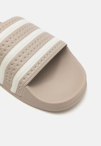 Sandal sa ravnim đonom od mekog bež gume s teksturiranim remenom koji ima tri bijele pruge. Ravni đon s glatkom završnom obradom i zaobljenim rubovima.
