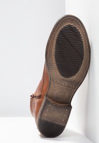 Semelle d'une botte en cuir marron usée avec une semelle en caoutchouc noir, indiquant la taille 37, posée debout contre un mur blanc en angle.