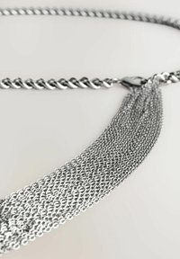 Ceinture en chaîne métallique argentée avec un design torsadé, ornée d'une frange de chaînes plus petites qui se cascade. Texture lisse, style contemporain.