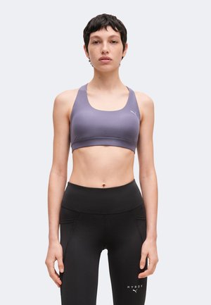 Mujer con el pelo corto y oscuro lleva un sujetador deportivo morado y unas mallas negras de cintura alta, de pie mirando al frente ante un fondo liso y claro.