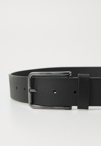 Zwarte leren riem met een gestructureerde afwerking. Voorzien van een glanzende zilveren gesp en een lus, met zichtbare gaatjes voor aanpassing.