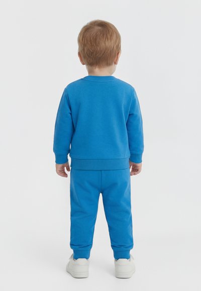 Tout-petit vu de dos portant un sweat-shirt bleu à manches longues, un pantalon assorti et des baskets blanches sur un fond uni.