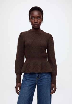 Femme aux cheveux courts portant un pull marron à manches longues avec un ourlet peplum et un jean bleu, debout devant un fond blanc uni.
