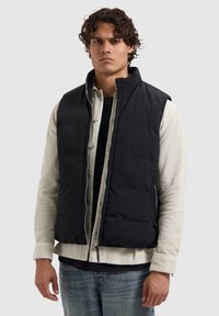 Zwarte mouwloze gewatteerde vest met een gestructureerd oppervlak, kraag en rits aan de voorkant. Draag het over een lichtgekleurde overhemd met knopen.