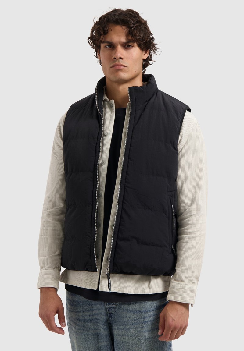 Zwarte mouwloze gewatteerde vest met een gestructureerd oppervlak, kraag en rits aan de voorkant. Draag het over een lichtgekleurde overhemd met knopen.