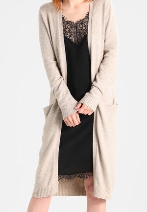 Femme portant une robe nuisette noire à bordure en dentelle sous un long cardigan beige avec poches, les mains jointes à la taille, jambes partiellement visibles.