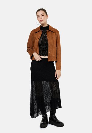 Vrouw draagt een bruine suède jas over een zwart kanten topje en een lange zwarte kanten rok, gecombineerd met zwarte chunky laarzen en oorringen.