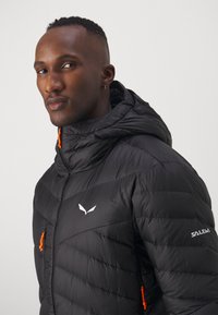 Giacca a vento nera con texture a trapuntatura orizzontale, dettagli arancioni sulle zip e logo bianco sul petto e sulla manica. Design con cappuccio.