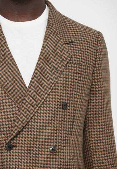Brun och svart houndstooth-mönstrad dubbelknäppt blazer med spetsiga slag och svarta knappar. Bärs över en vit skjorta.
