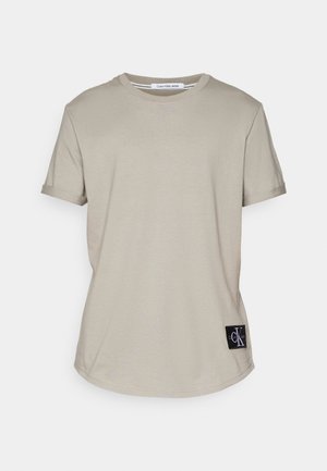 Beige katoenen t-shirt met een ronde hals en opgerolde korte mouwen. Heeft een zwart label met wit logo aan de onderkant. Soepele textuur.
