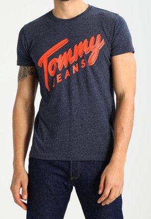 Tummansininen t-paita, joka on valmistettu puuvillasekoitteesta ja jossa on lyhyet hihat. Esittelee suurta punaista "Tommy Jeans" -logoa voimakkaassa, vinossa fontissa.