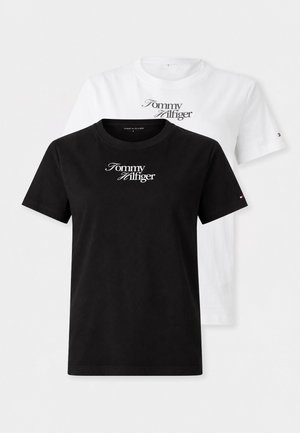 Femme portant un t-shirt blanc Tommy Hilfiger, un pantalon beige avec une ceinture marron, des mocassins noirs et tenant un sac à main noir avec des lanières blanches.