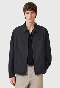 Zwarte lichte jack met een kraag, ritssluiting aan de voorkant en een geribbeld textuur. G gedragen over een donker overhemd, met beige broeken. Minimalistisch ontwerp.