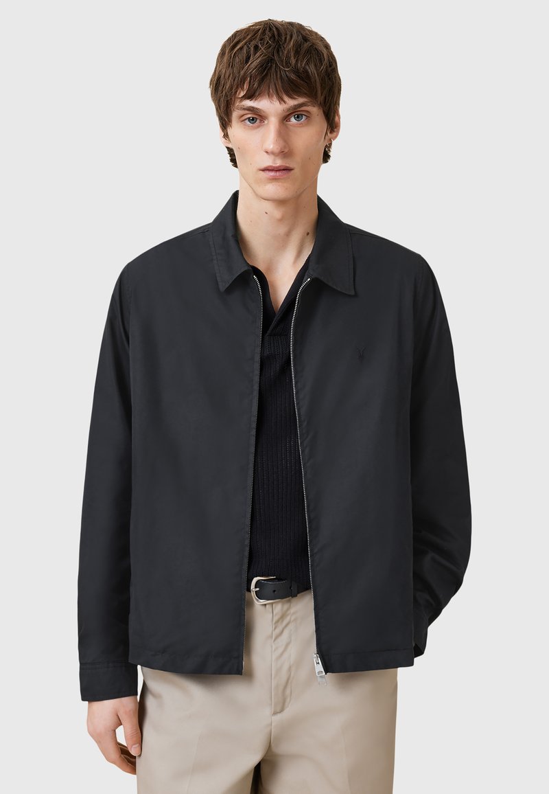 Zwarte lichte jack met een kraag, ritssluiting aan de voorkant en een geribbeld textuur. G gedragen over een donker overhemd, met beige broeken. Minimalistisch ontwerp.